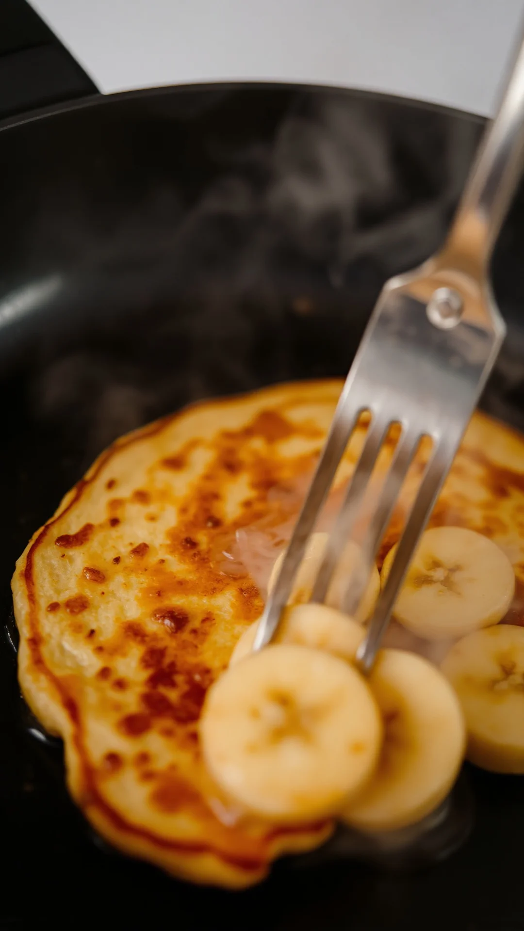 Close-up macro shot of freshly cooked banana pancake being flipped in non-stick pan, golden brown surface, steam rising, prof