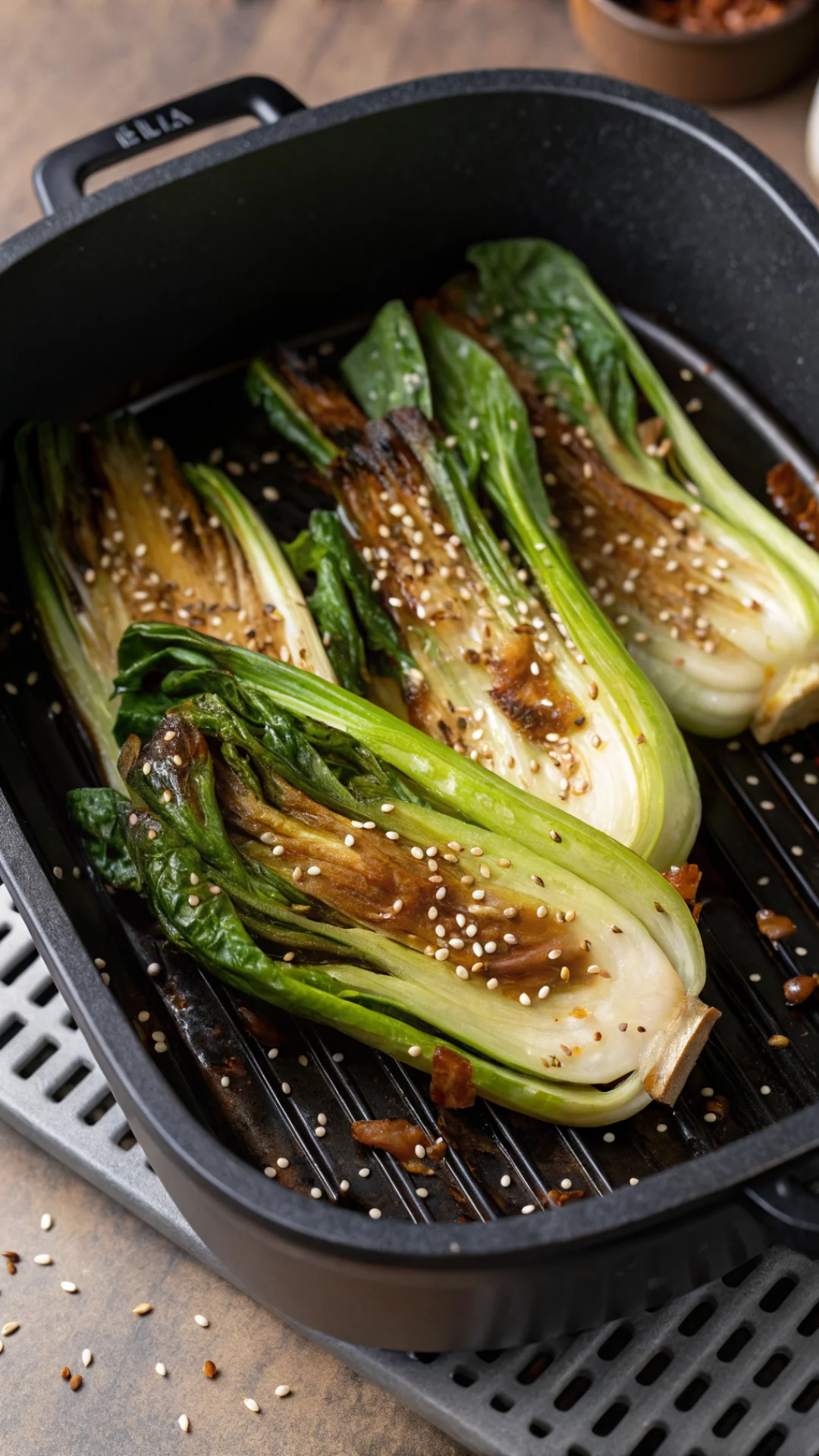 Close-up overhead view of crispy brown pak choi halves in air fryer basket, glistening with glaze, sesame seeds sprinkled on